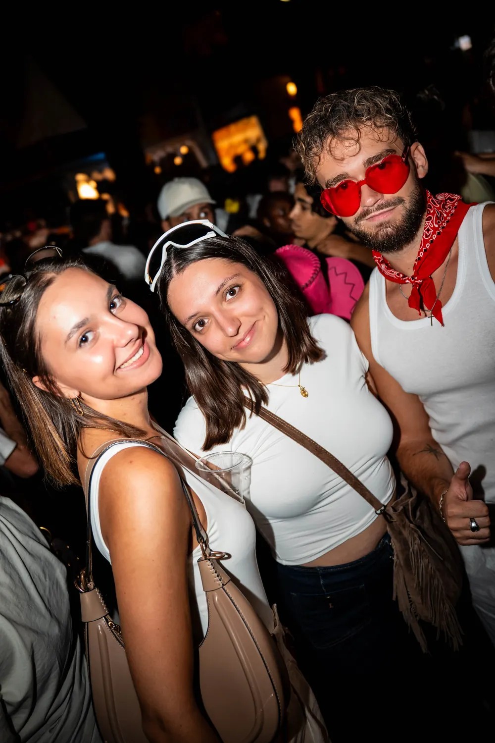 Trois amis en tenue de feria, lunettes cœur rouges et bandana, au milieu de la foule de nuit